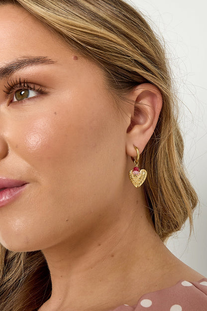 Close portrait of woman wearing gold heart drop earrings with pink and red beads, styled with light makeup and polka dot top