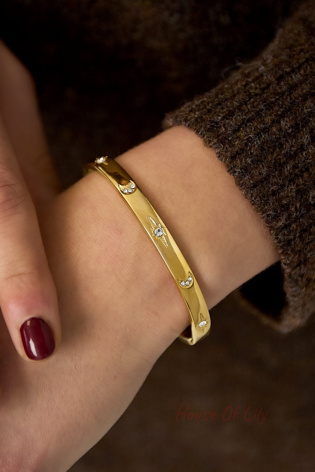 Close-up of gold-tone crystal accent bangle bracelet worn on wrist with brown knit sleeve and dark red nail polish