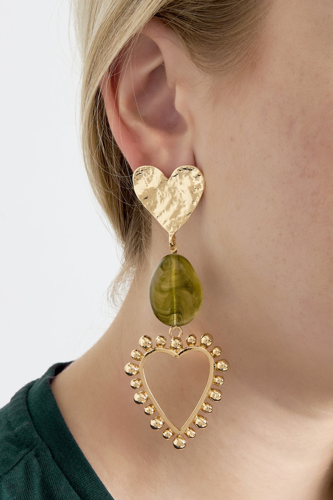 Close-up of a woman wearing heart drop earrings featuring hammered gold heart studs, olive green marbled resin bead, and gold beaded open heart frame