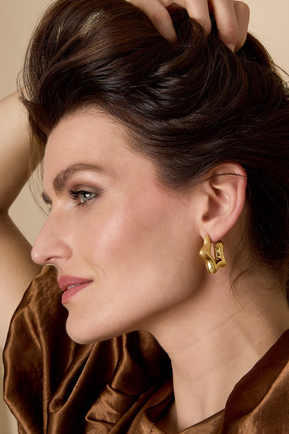 Close-up of woman wearing gold sculptural hoop earrings, showing their playful organic wave shape paired with a brown velvet outfit