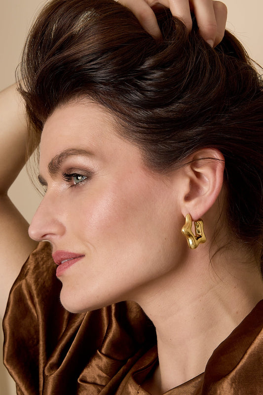 Close-up of woman wearing gold sculptural hoop earrings, showing their playful organic wave shape paired with a brown velvet outfit