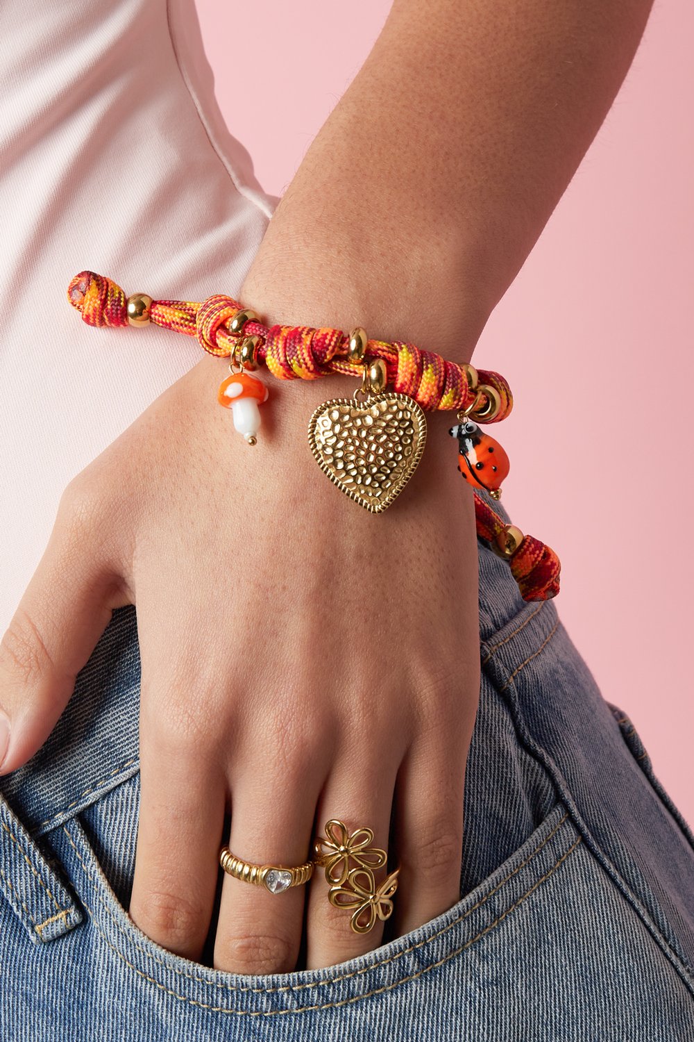 Playful orange and yellow rope knot bracelet with gold heart, mushroom, and ladybug charms on a person's wrist.