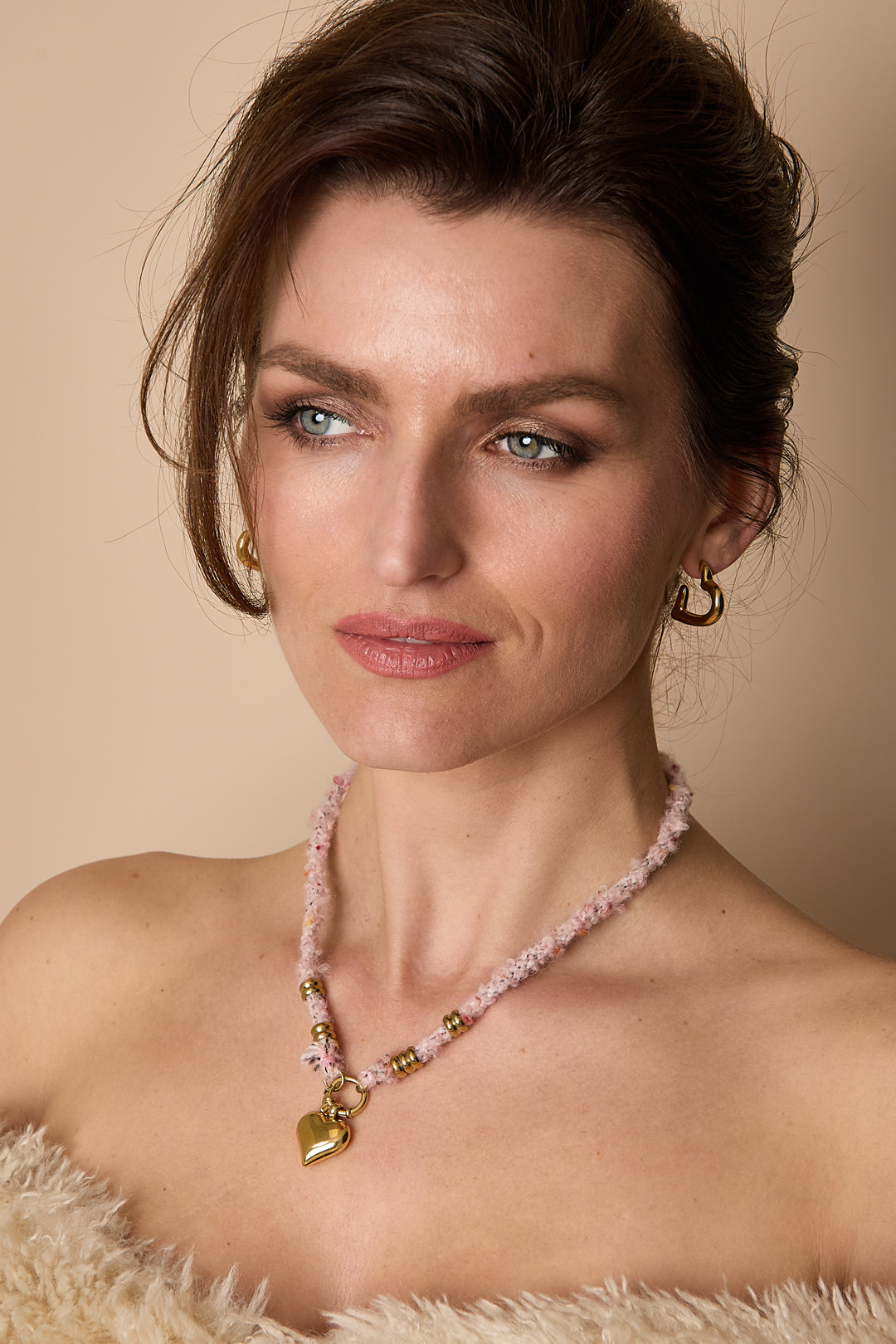 Woman wearing a soft pink wool rope necklace with gold rings and shiny gold heart pendant, styled with neutral makeup and light fur top