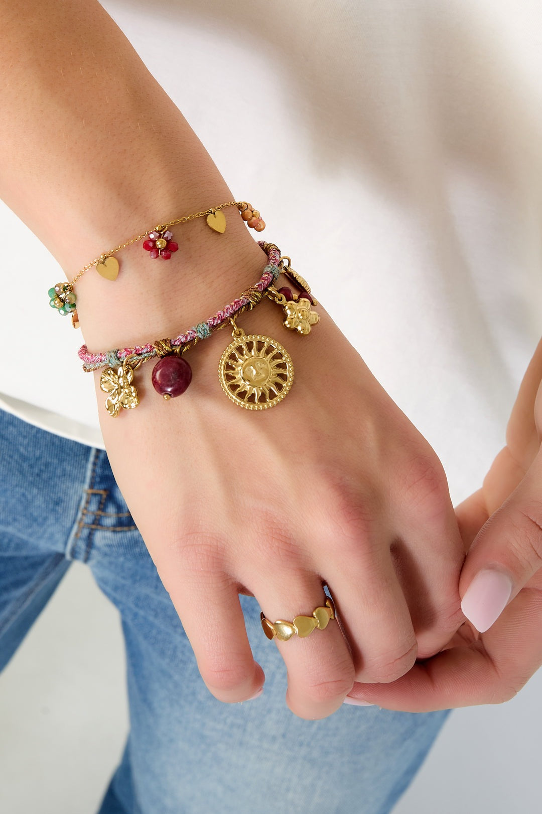 Close-up of woman wearing braided charm bracelet with gold-tone sun and flower charms, paired with gold heart ring and floral charm bracelet