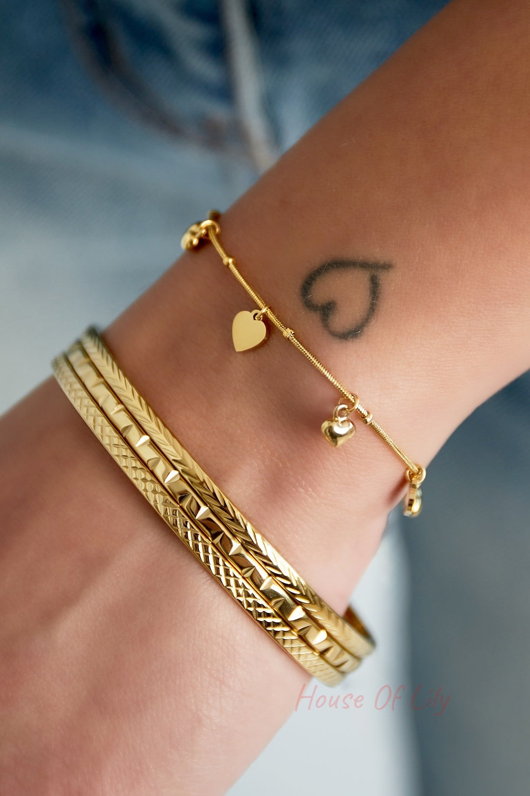 Close-up of a wrist wearing two textured gold bangles and a delicate gold charm bracelet against denim