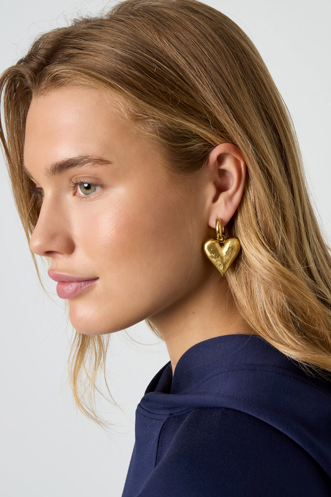 Blonde woman wearing gold heart charm drop earring with a navy top, side profile
