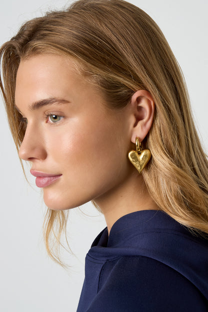 Blonde woman wearing gold heart charm drop earring with a navy top, side profile