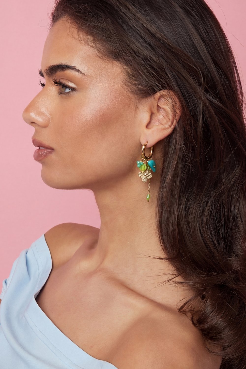 Gold hoop earrings featuring whimsical hanging charms in turquoise and lime green on a woman against a pink background.