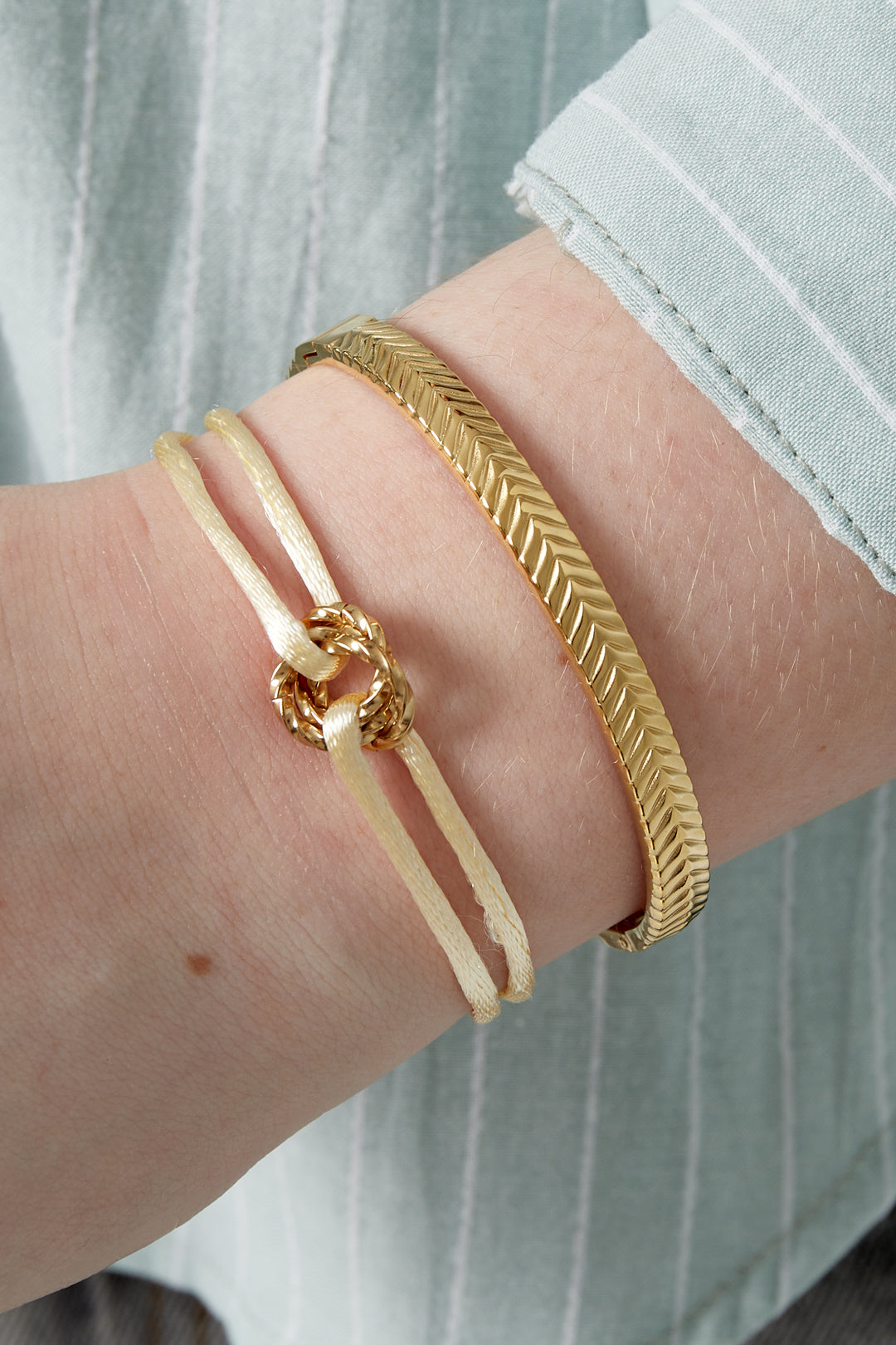 Cream knot charm bracelet and gold bracelet on woman's wrist wearing green striped shirt