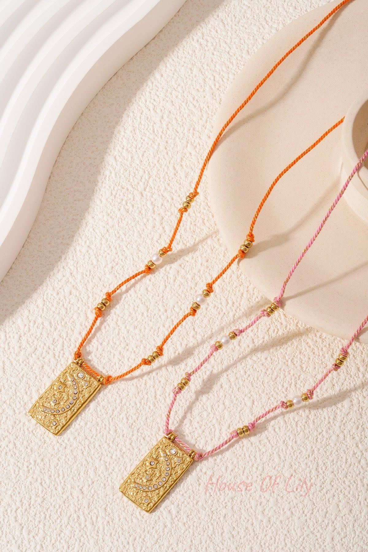 Two braided moon pendant necklaces in orange and pink laid on textured surface