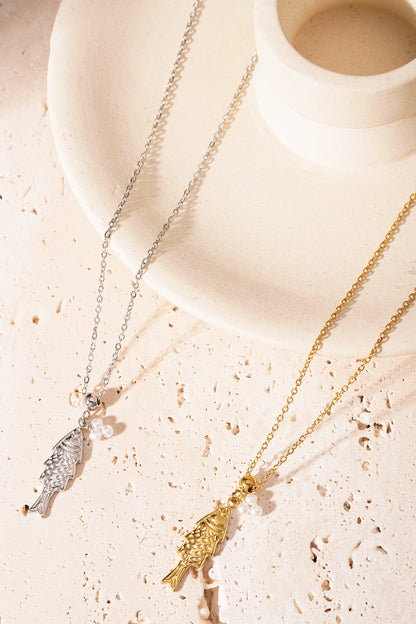 Silver and gold fish pendant necklaces laying on a cream textured surface beside a cream candle holder