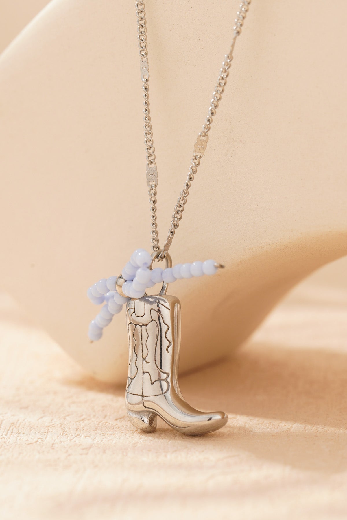 Silver-tone cowboy boot pendant necklace with pale blue beaded bow displayed on black card