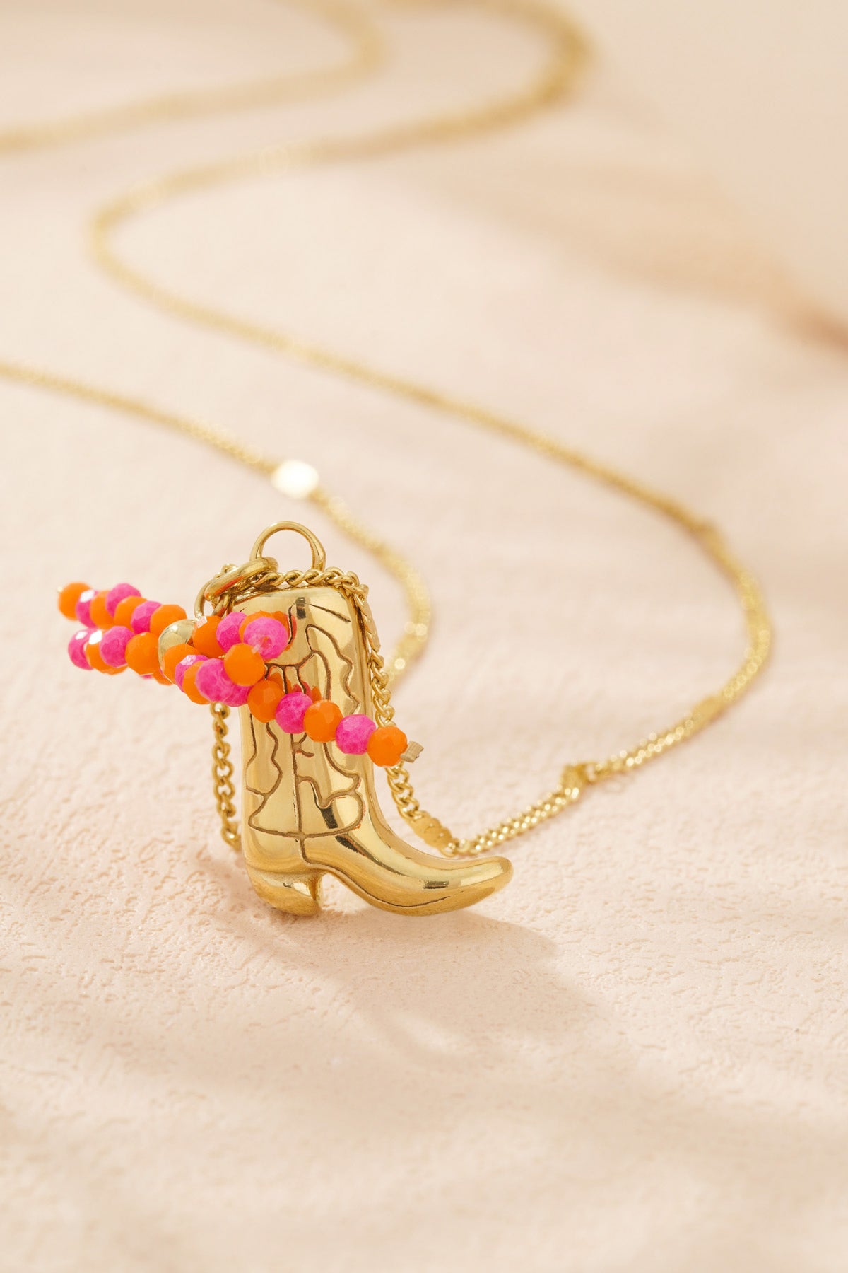 Close-up of gold-tone cowboy boot pendant necklace with pink and orange beads on cream background
