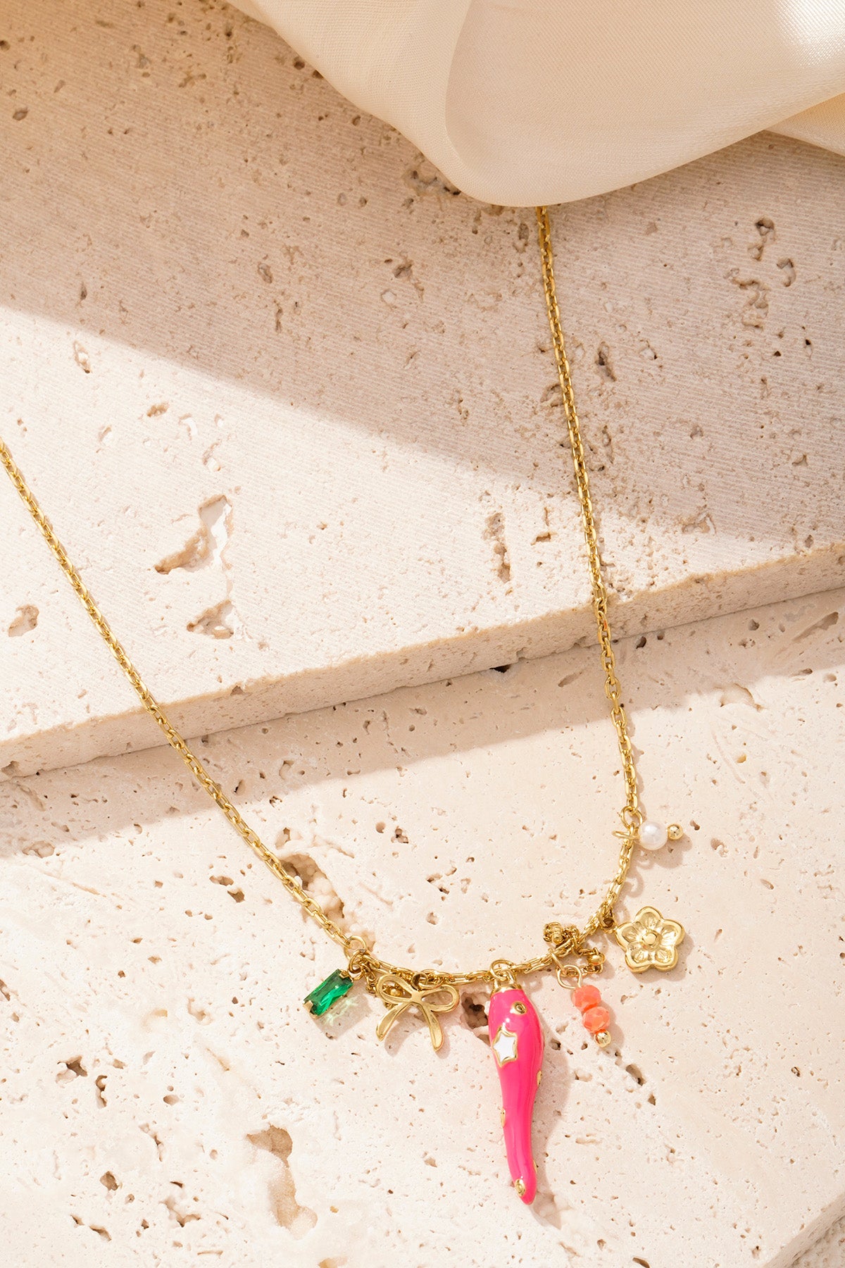 Close-up of gold-tone charm necklace with pink pepper, flower, bow and beads displayed on textured stone surface