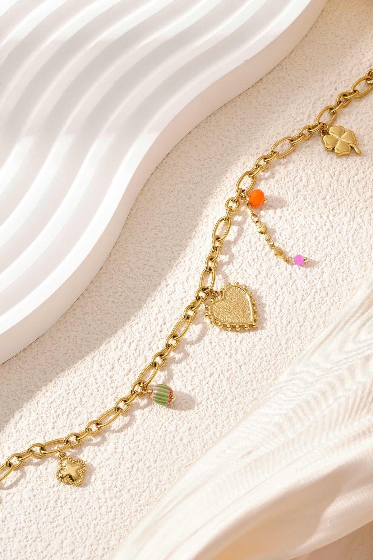 Artistic image of golden charm link necklace showing the charm arrangement on a wavy white surface with delicate fabric.