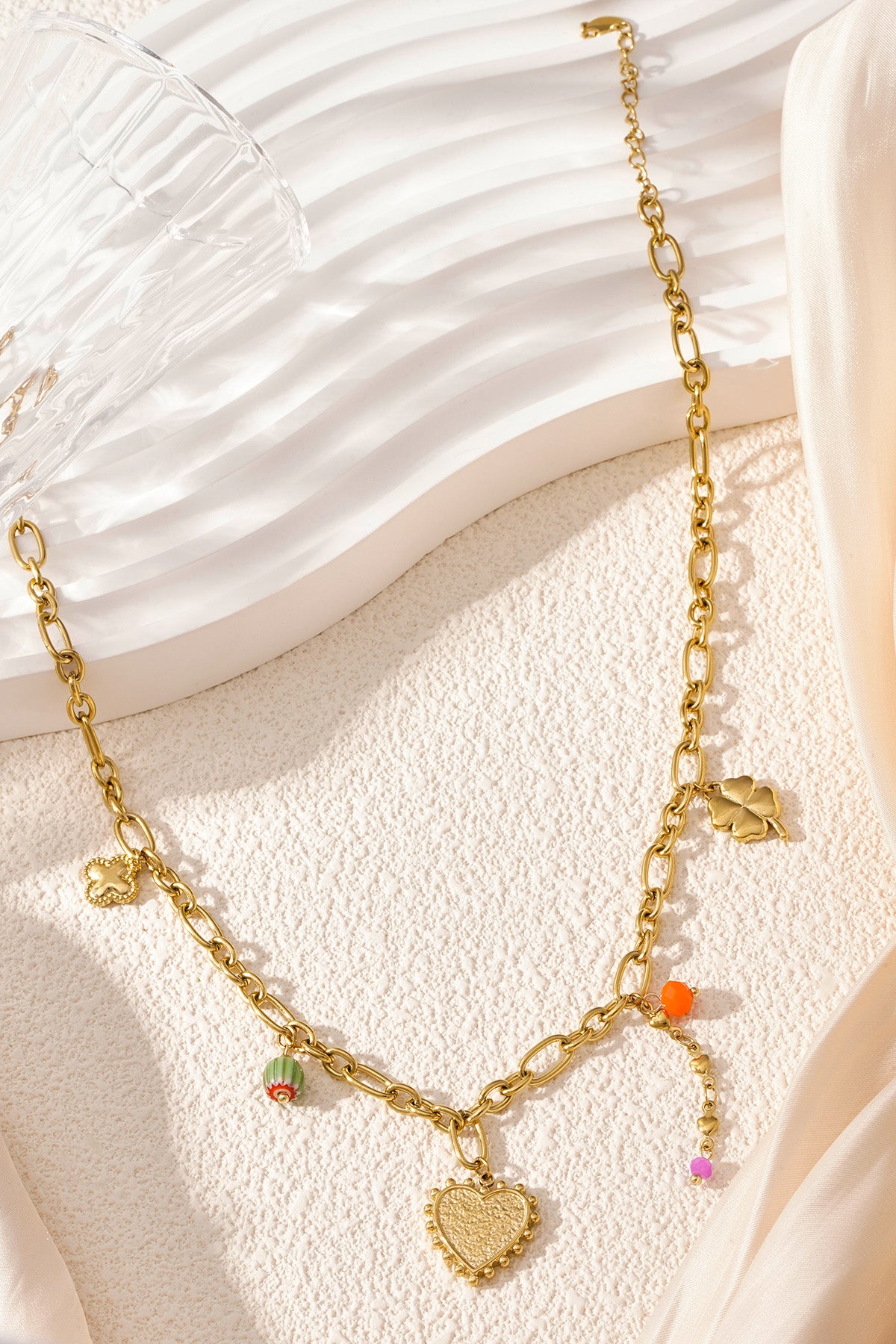 Detailed product shot of golden charm link necklace arranged on a light textured background with a glass, showing the shine of the gold and colourful charms.