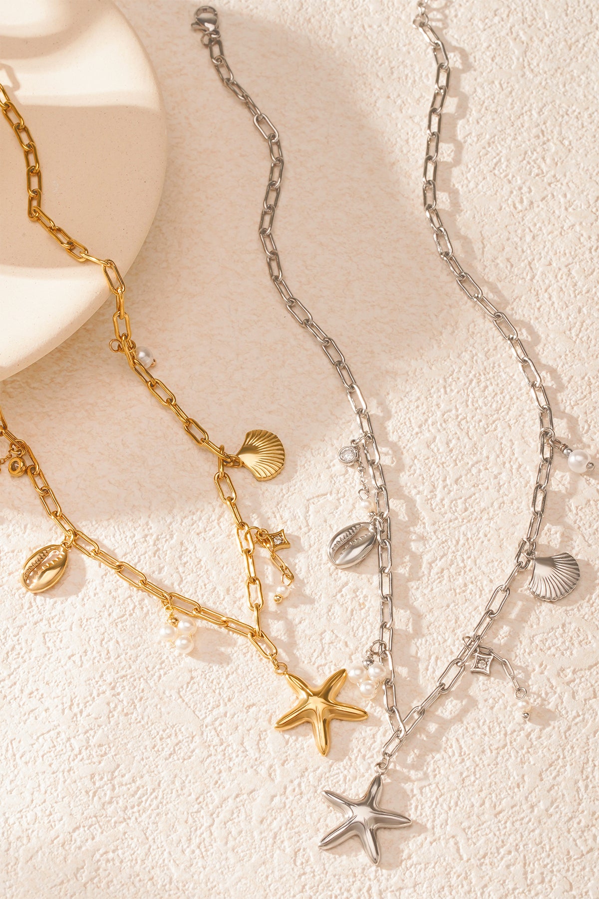 Gold and silver seaside charm necklaces laid flat on a textured white surface, showcasing starfish, shells and pearl clusters