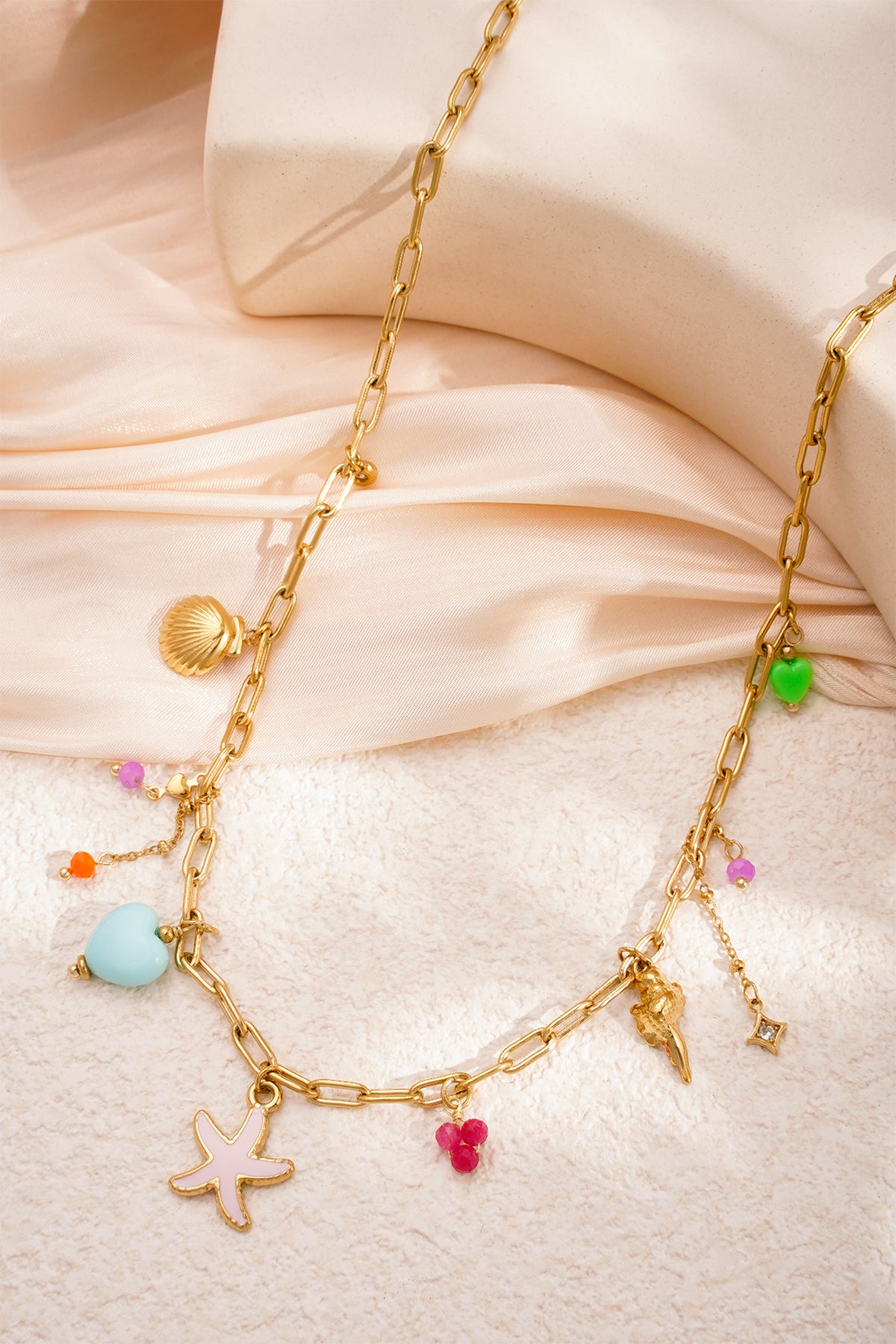 Gold-tone charm necklace with marine-themed pendants and pastel beads styled on beige fabric and textured surface