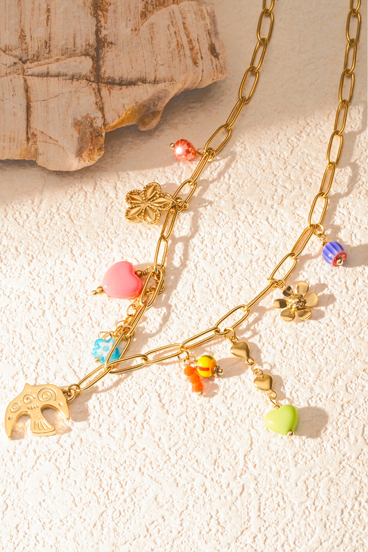 Soft natural light on gold chain necklace with bright playful charms arranged on textured cream surface