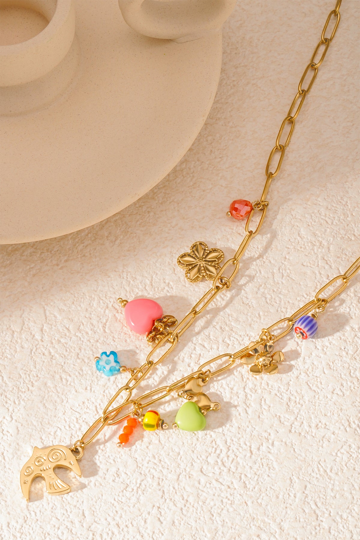 Close-up of necklace laid on textured surface highlighting colourful heart, flower, bird and bead charms on gold links