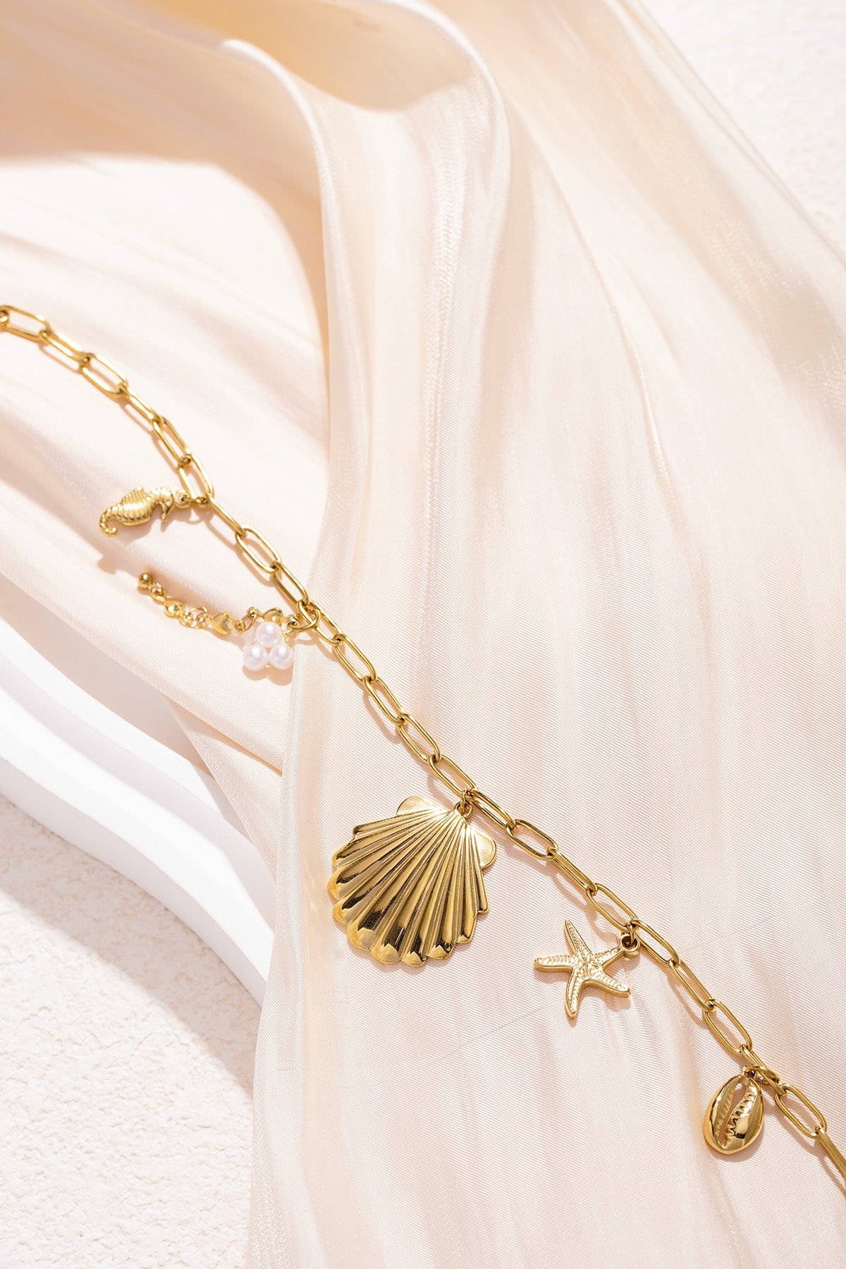Detailed view of the gold seaside charm necklace with links and charms lying on light-coloured background showing pearl cluster and cowrie shell.