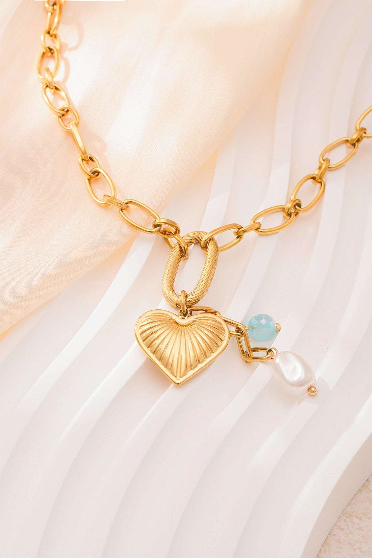 Close-up of gold textured heart pendant necklace featuring aqua bead and pearl on soft cream fabric