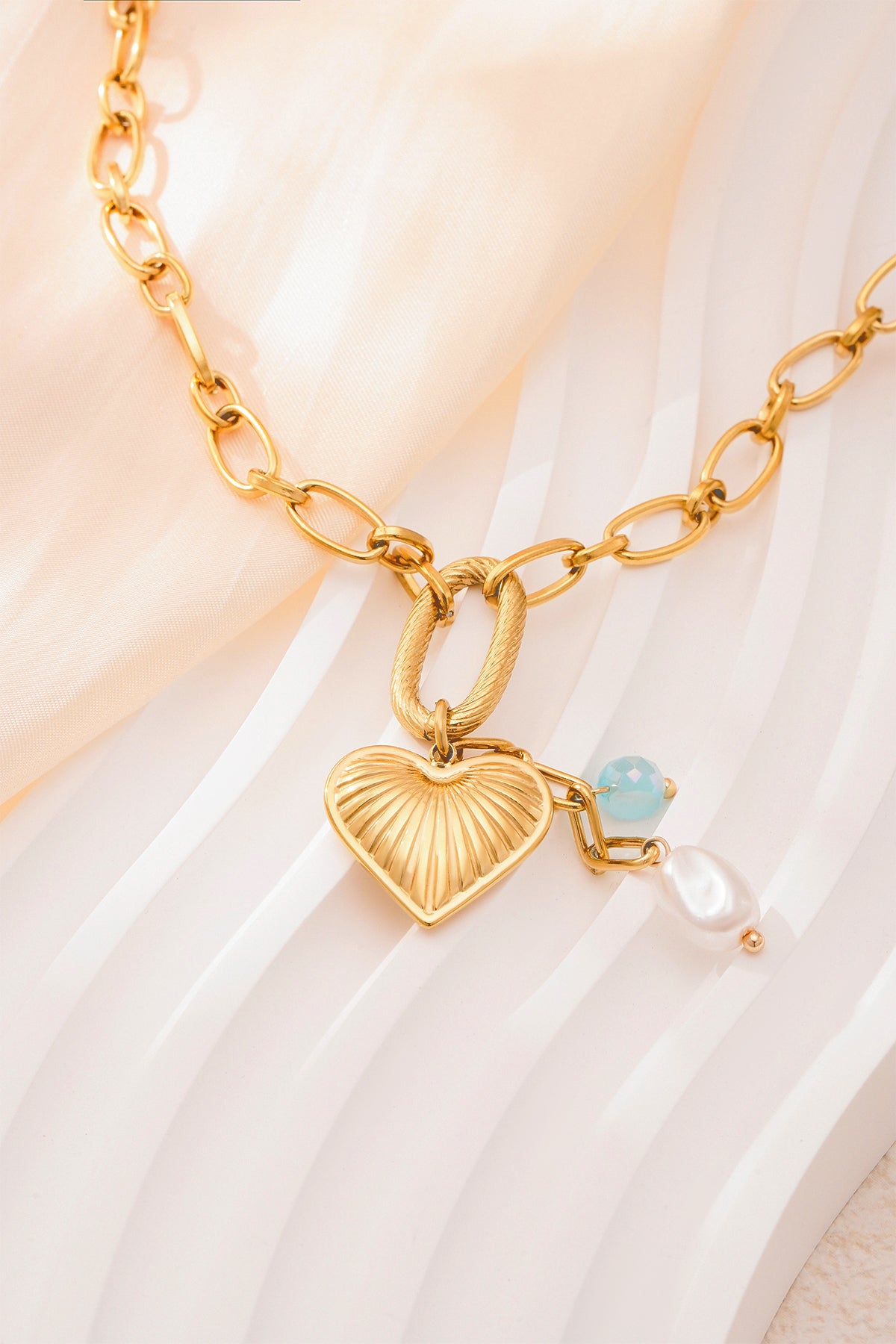 Close-up of gold textured heart pendant necklace featuring aqua bead and pearl on soft cream fabric