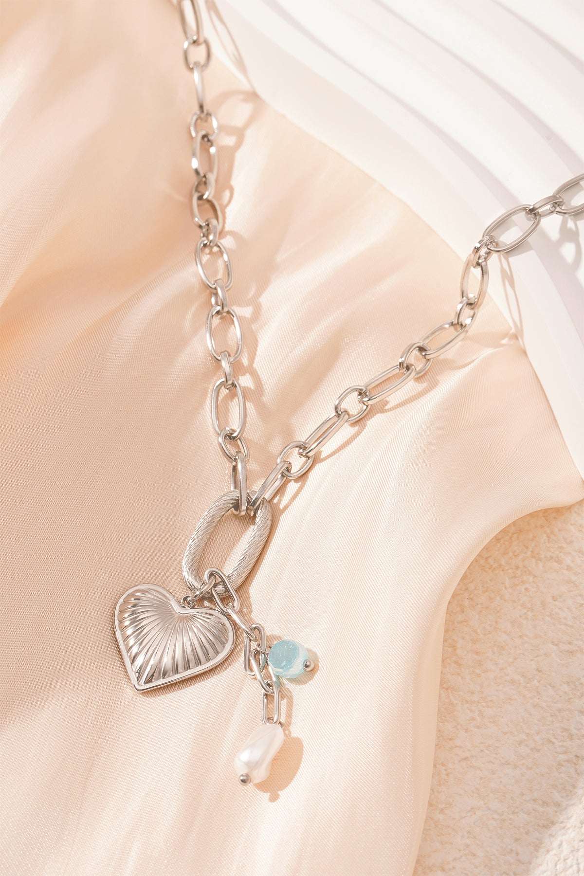 Close-up of silver version of chain necklace with heart pendant, aqua bead and pearl on cream fabric