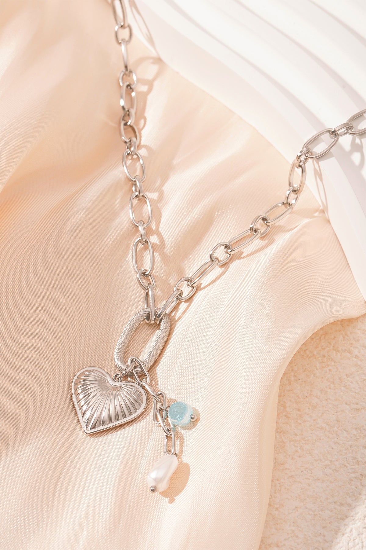 Close-up of silver version of chain necklace with heart pendant, aqua bead and pearl on cream fabric