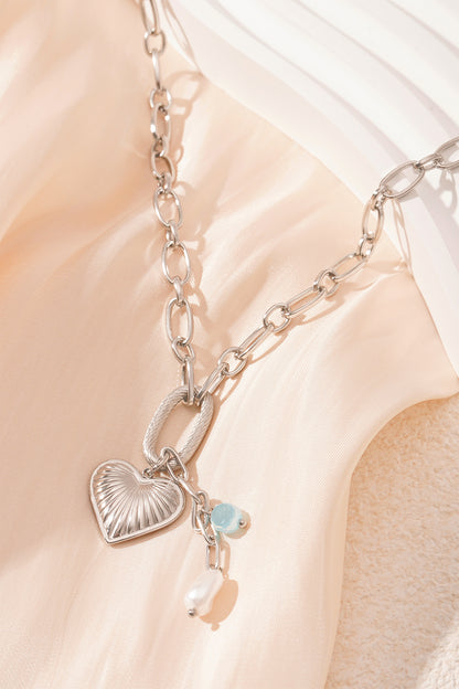 Close-up of silver version of chain necklace with heart pendant, aqua bead and pearl on cream fabric
