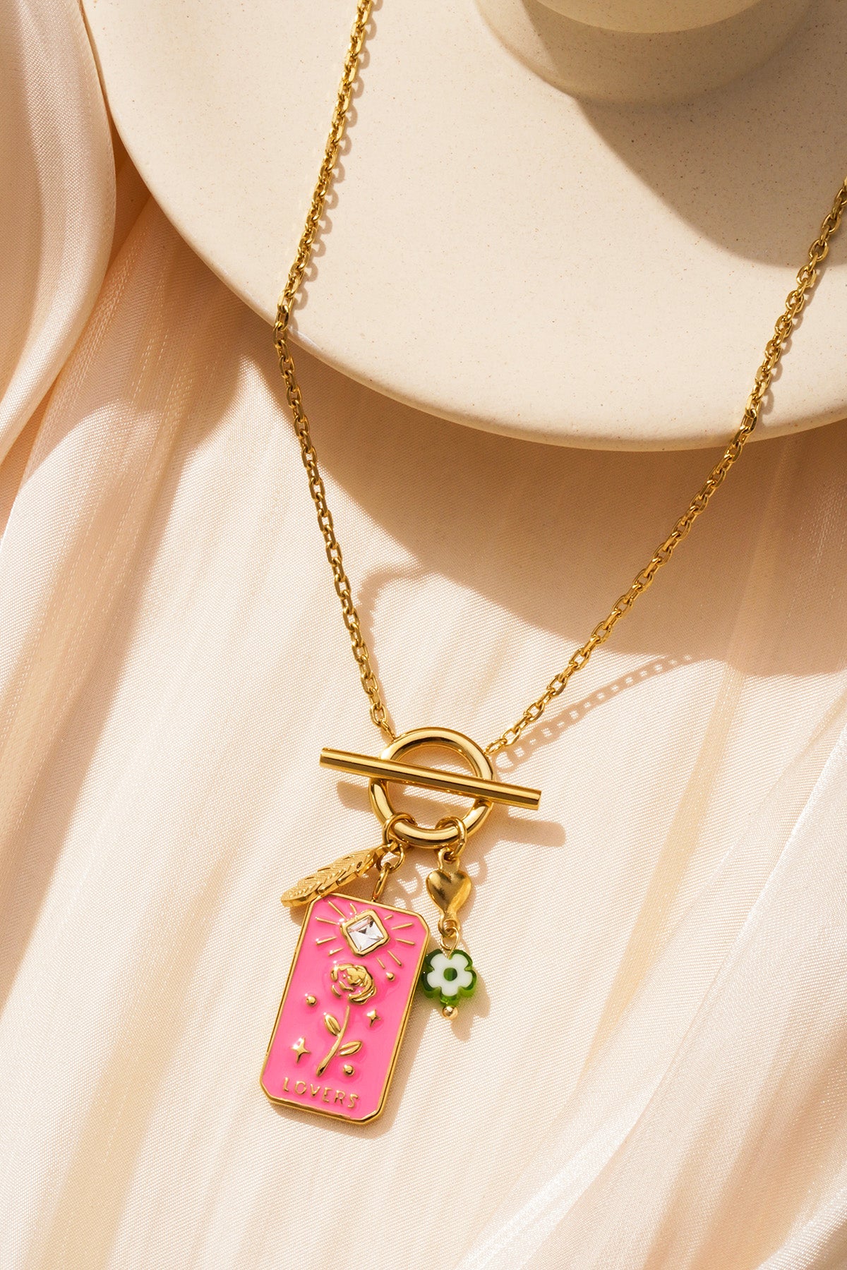 Close-up of the necklace charms with pink floral tag and gold toggle clasp on soft beige fabric background