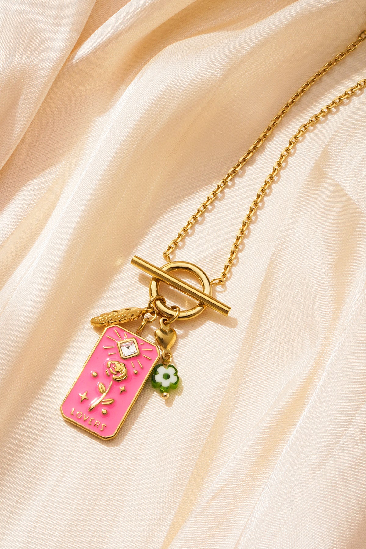Gold-tone pendant necklace with pink floral tag and charms laid on shimmering beige fabric