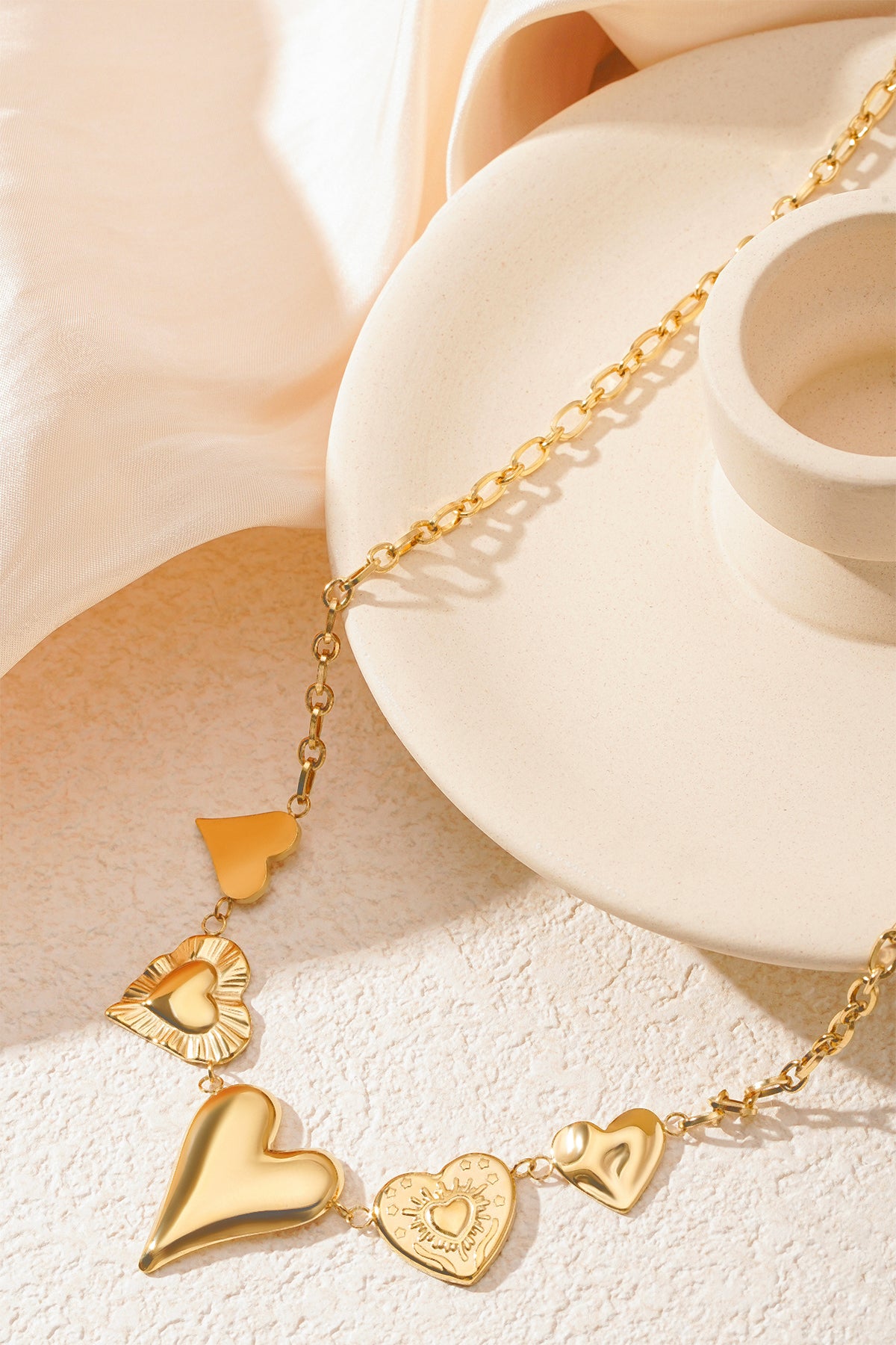 Gold multi heart charm necklace positioned on a beige ceramic dish with soft fabric background