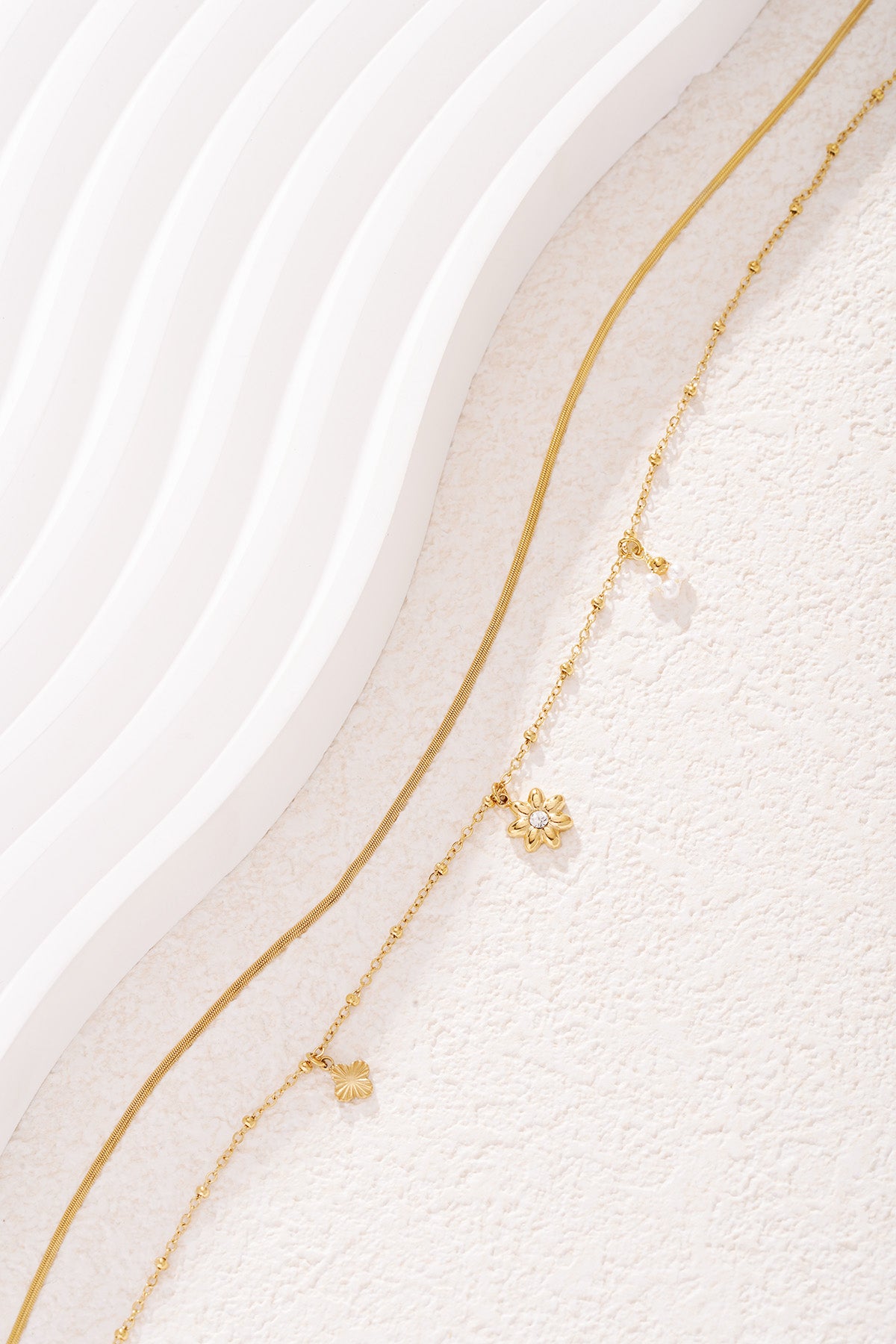 Gold layered necklace with flower, pearl charm details close-up on white textured background