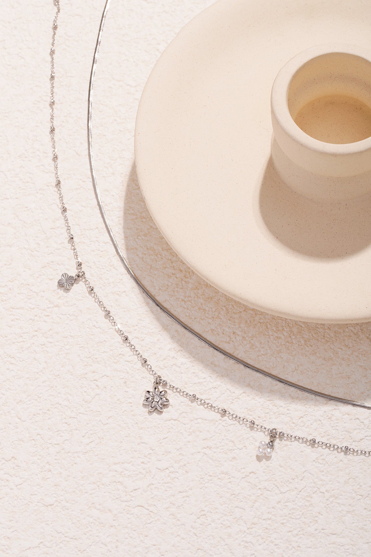 Silver layered necklace with flower and pearl charms detail with beige ceramic cup on textured background