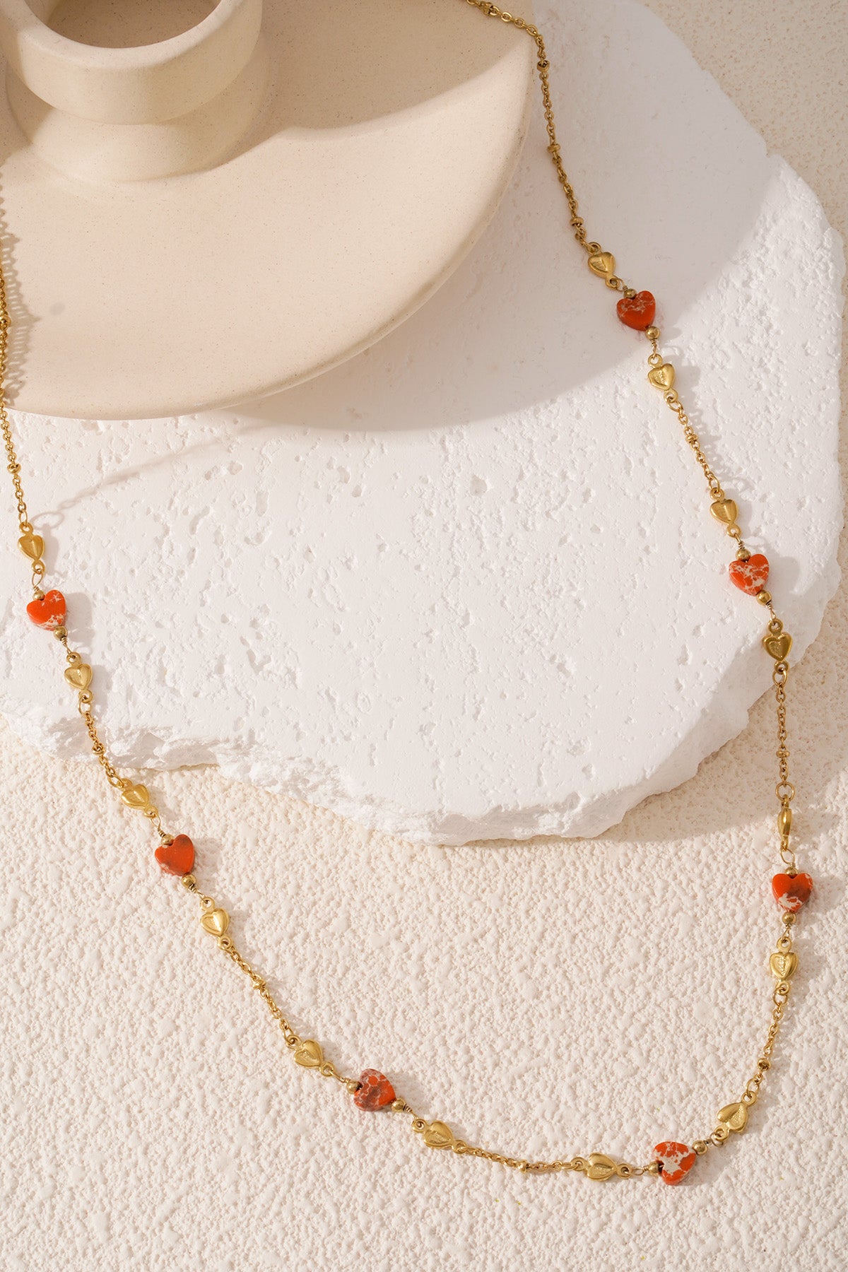 Gold-tone chain necklace with orange and gold heart-shaped beads elegantly displayed on textured white surface