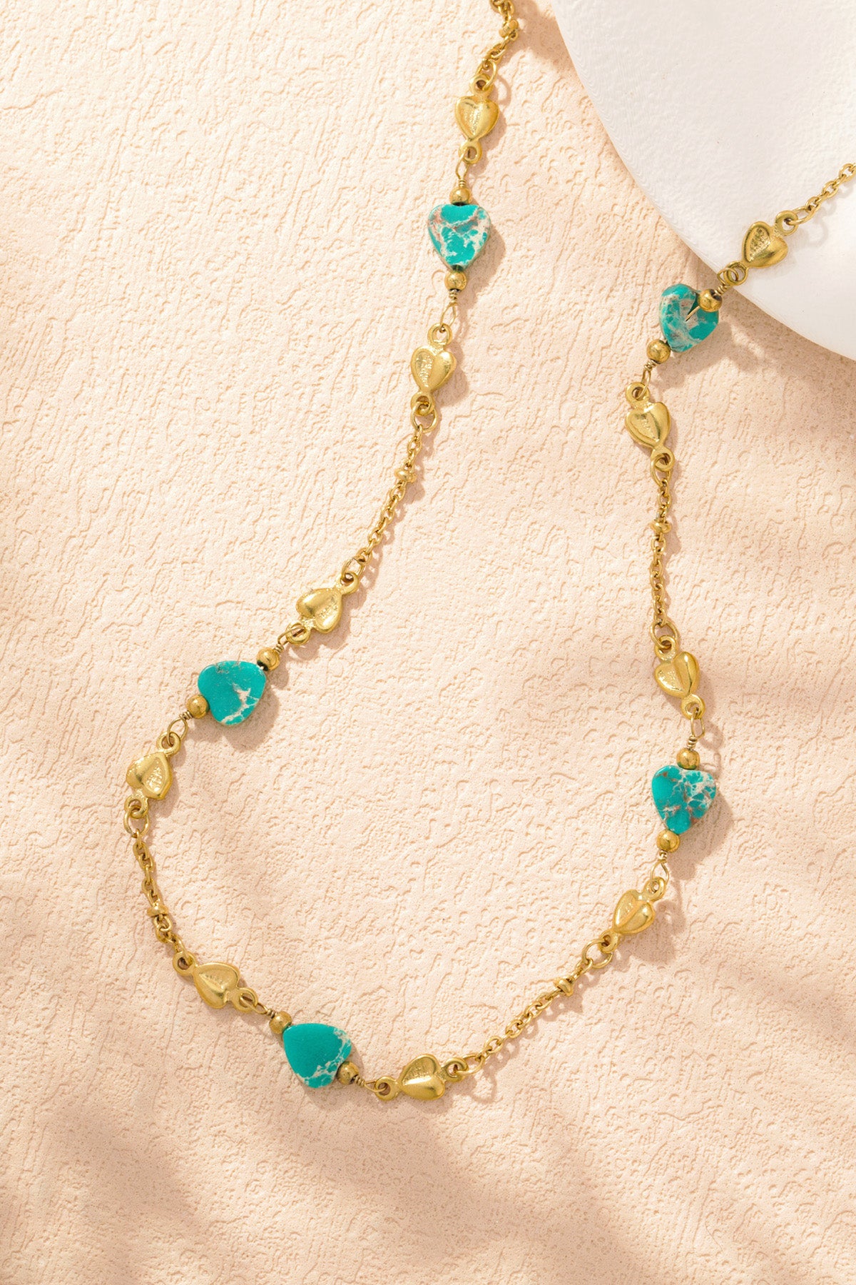 Close-up of gold-tone chain necklace with turquoise and gold heart-shaped beads on white textured surface