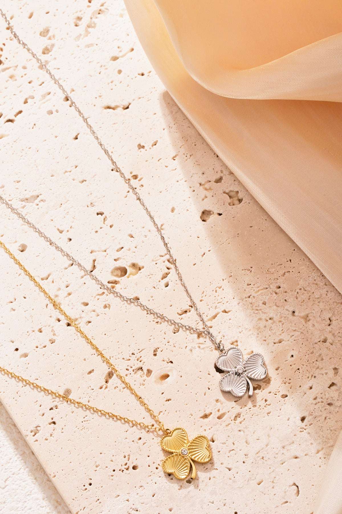 Gold and silver clover necklaces arranged on textured stone surface with a soft peach coloured fabric softly draped