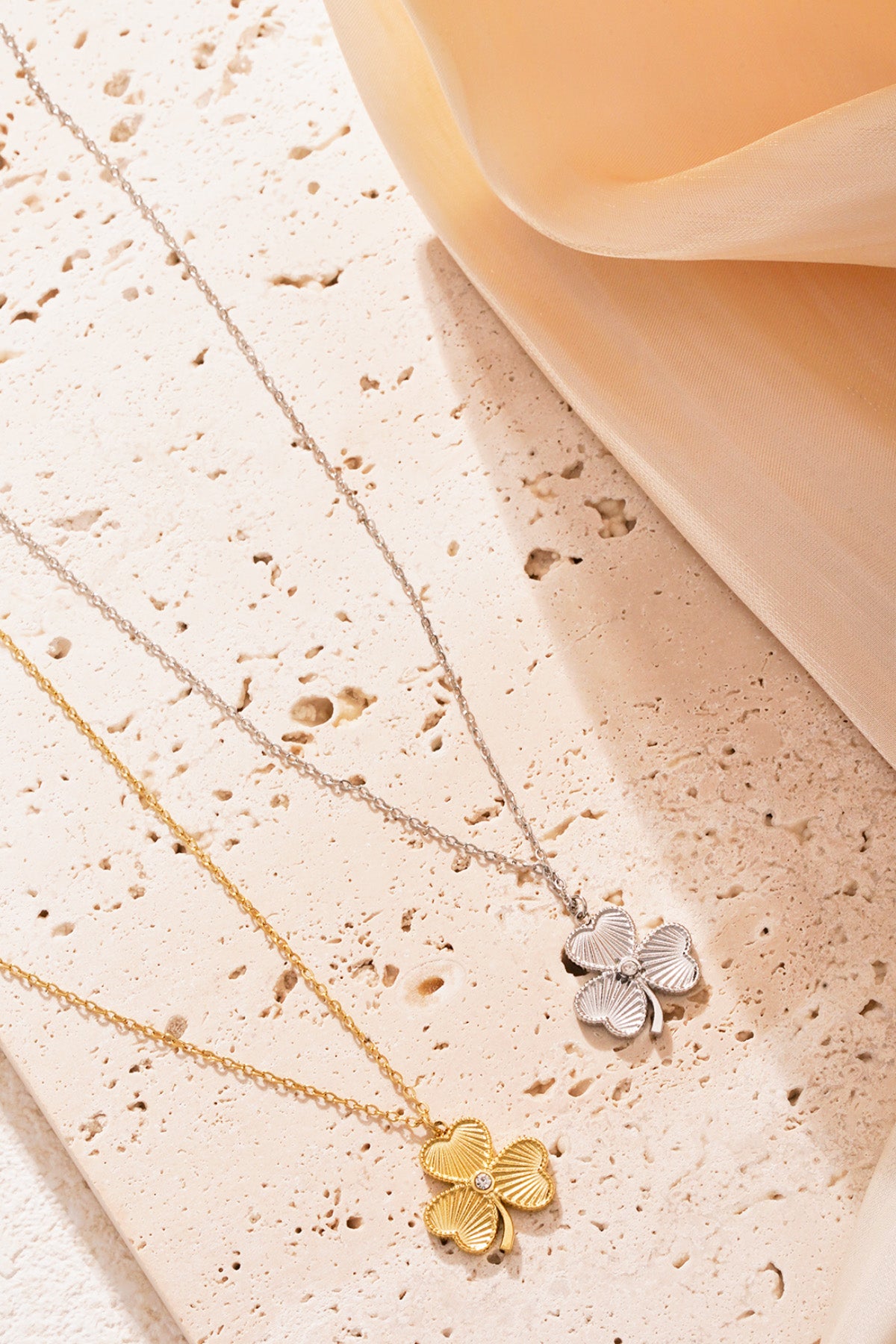 Gold and silver clover necklaces arranged on textured stone surface with a soft peach coloured fabric softly draped