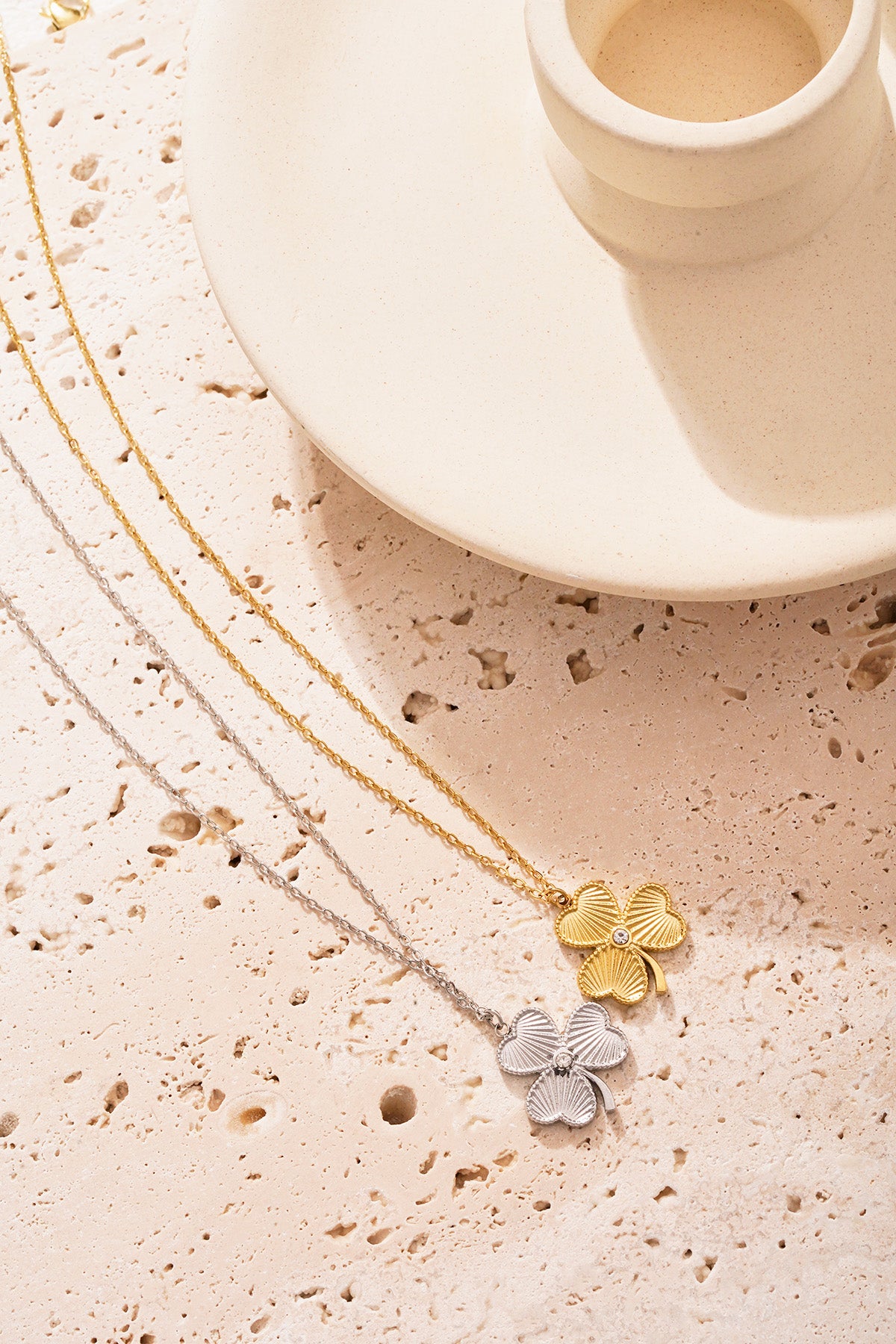 Gold and silver clover necklaces displayed on textured stone surface beside off-white ceramic holder