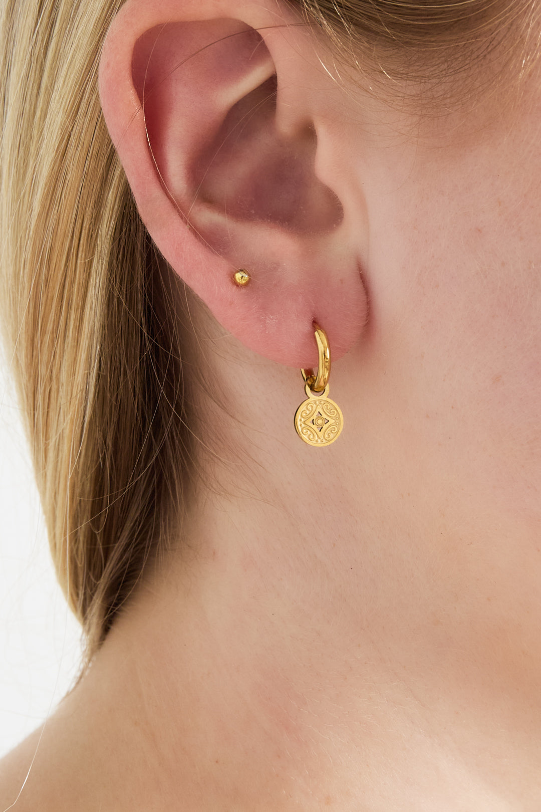 Close-up of woman's ear wearing gold-tone hoop earring with round engraved charm, showing youthful and minimal look