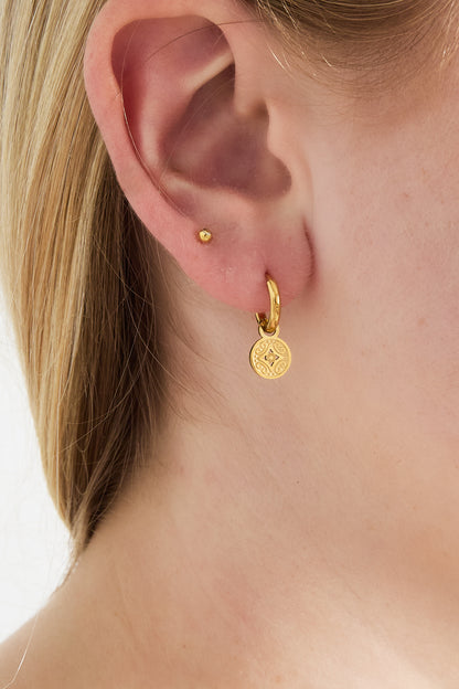 Close-up of woman's ear wearing gold-tone hoop earring with round engraved charm, showing youthful and minimal look