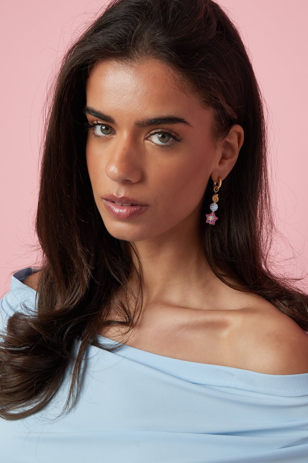Whimsical gold dangle earrings featuring a blue striped bead and a pink starfish charm on a woman against a pink wall.