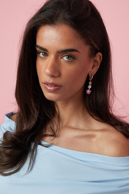 Whimsical gold dangle earrings featuring a blue striped bead and a pink starfish charm on a woman against a pink wall.