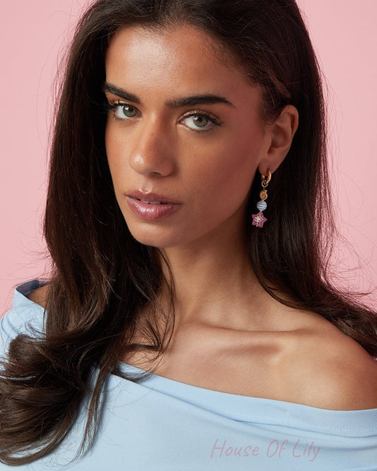 Whimsical gold dangle earrings featuring a blue striped bead and a pink starfish charm on a woman against a pink wall.