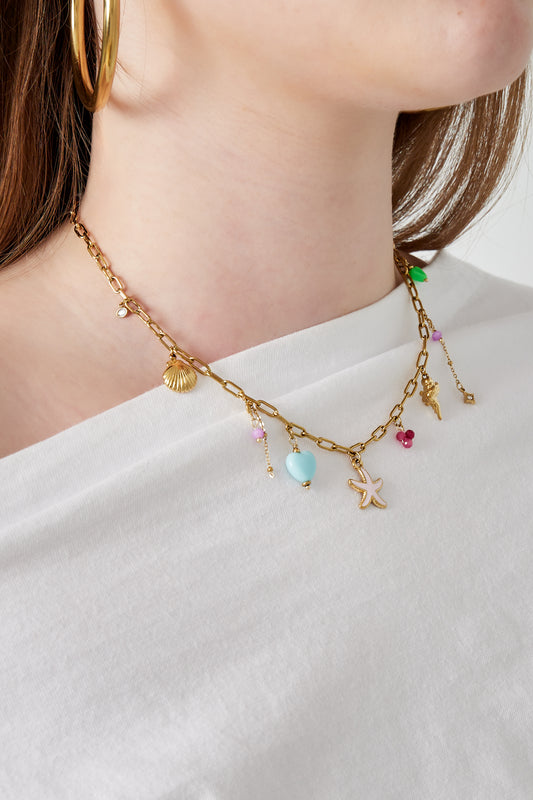 Close-up of woman wearing gold-tone charm necklace with pastel starfish and seashell charms on white top
