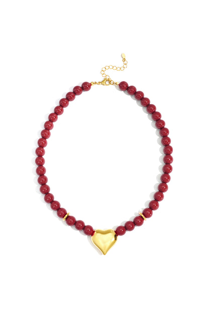 Close-up of glossy crimson red beaded necklace with shiny gold heart pendant and gold lobster clasp on white background