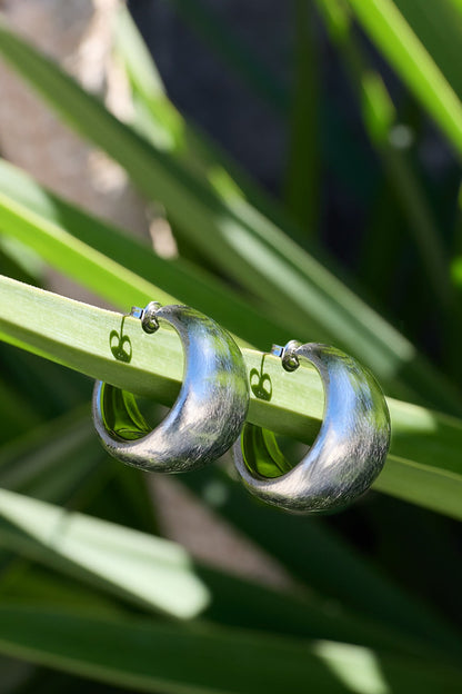 Chunky Aura Loops: Thick, brushed silver hoop earrings resting on a green leaf.