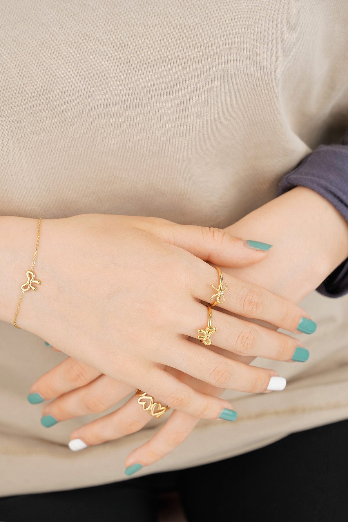 Hands showcasing multiple gold rings including bow rings and bracelet with teal and white nail polish