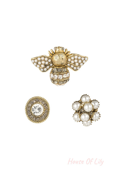 Set of three decorative women's brooches including a pearl and rhinestone bee, a circular crystal-encrusted brooch, and a pearl cluster flower on white background